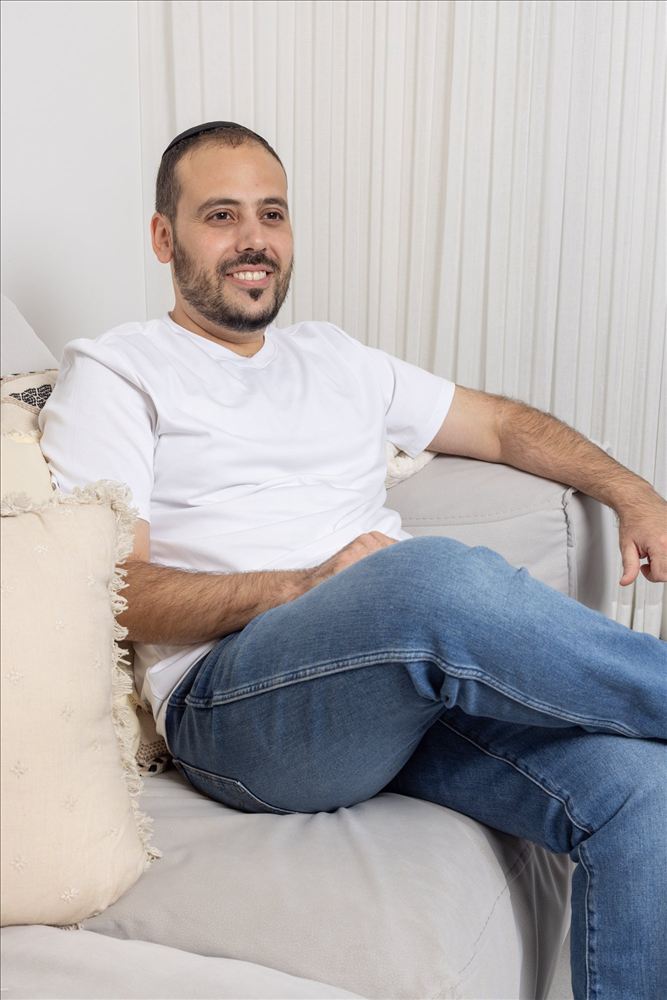 גבר בחולצה לבנה יושב בנינוחות על ספה ומחייך